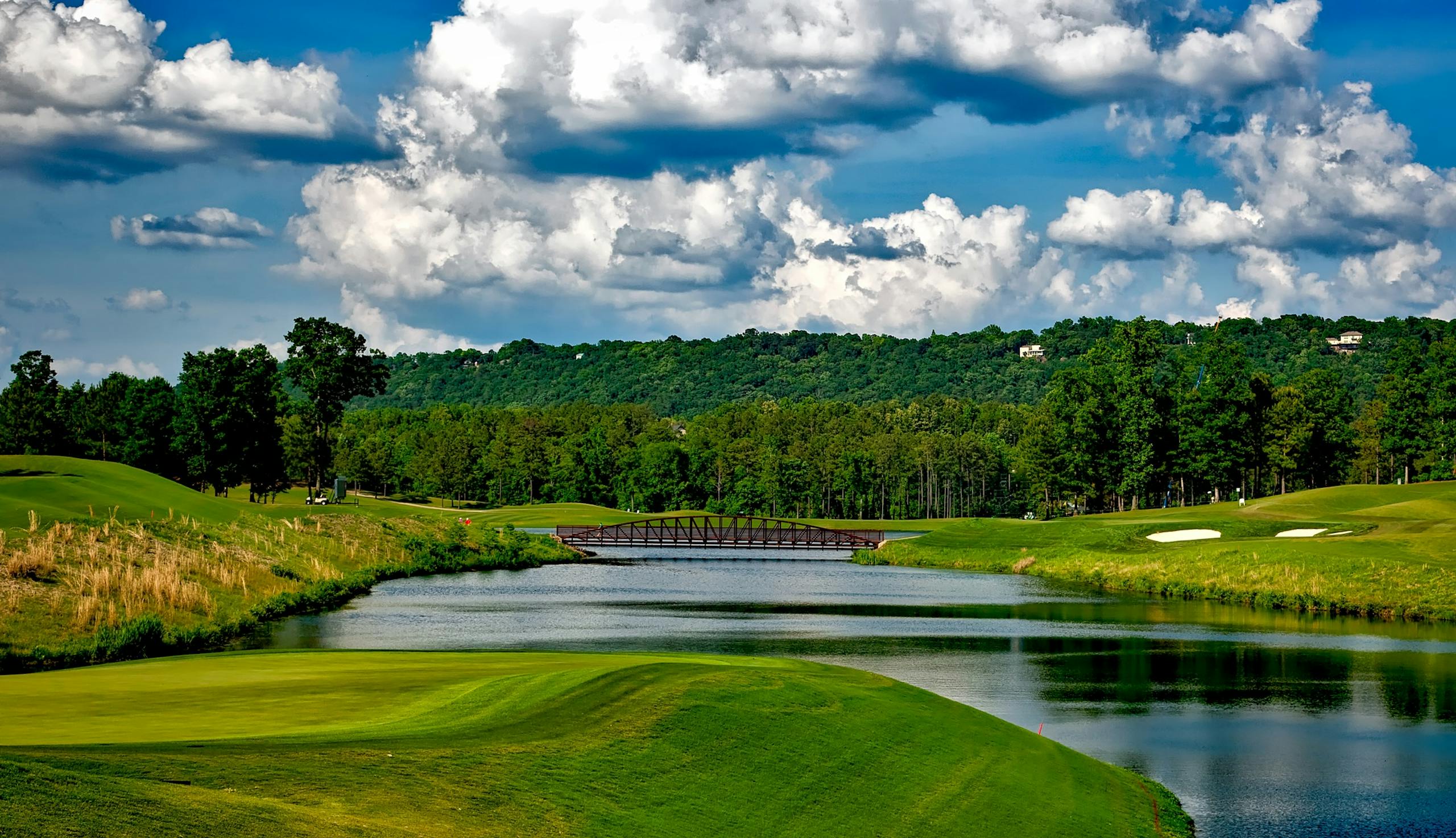 Scenic golf course with lush greenery, river, and a bridge under a bright summer sky.